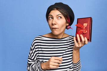 Adorable young woman in stylish beret and striped shirt posing isolated with New Year present, shaking box at her ear and looking up with pensive thoughtful facial expression, guessing what is inside