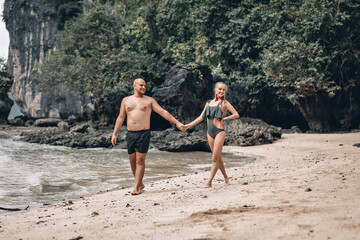 Attractive trendy young couple in white clothes are walking along on the beach and holding hands. Beautiful blonde with long hair and a bald man are on vacation. Phuket. Thailand..