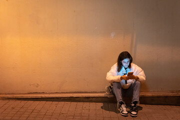 Beautiful young woman using her phone sitting in the night city street waiting for someone. Alone girl in the street at night. © deliris