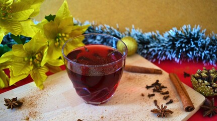 Spiced mulled wine surrounded by cinnamon sticks, cloves, star anise, a red reindeer, candy canes, tinsel in the shape of a fir branch, pins, a golden ball and golden poinsettias on a table 