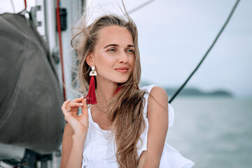 portrait of happy pretty girl with white dress, long red earrings and long curly blonde hair standing on yacht at summertime. looking at camera with toothy smile. close up