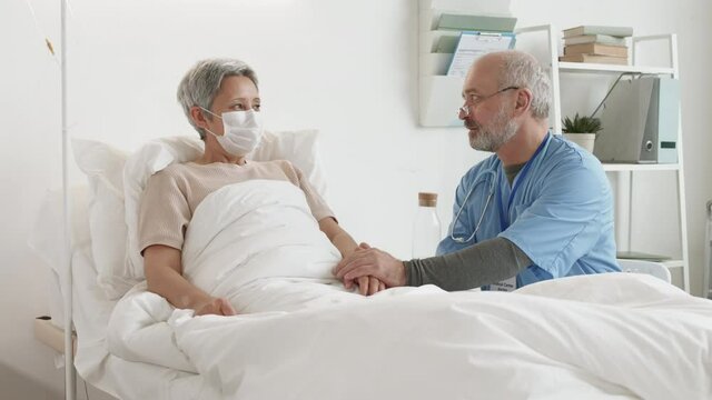 Lockdown of aged Caucasian male doctor sitting near bed in hospital ward and patting old Asian woman wearing medical mask on hand