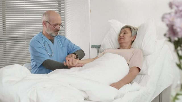 Medium shot of Caucasian physician wearing blue overall sitting on chair near bed and measuring pulse of ill old Asian woman lying on it