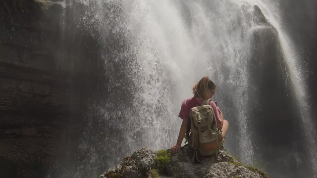 A girl hiking is sitting on a rock under a waterfall in a nature reserve. Young sporty woman takes a picture with the phone. Relaxing movement of water waves.  
Water is falling in the background.