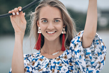 portrait of happy pretty girl with colorful dress,long red earrings and long curly blonde hair sitting on yacht at summertime. looking at camera with toothy smile. Close up photo.