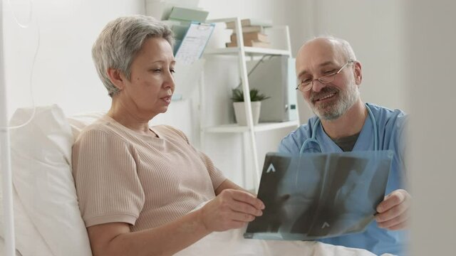 Medium shot of ill old Asian woman and aged Caucasian physician wearing blue overall analyzing x-ray picture in hospital ward