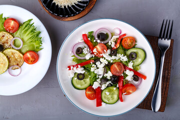 White plate with grilled zucchini with cherry tomatoes on lettuce leaves and greek salad on a concrete background near sesame seeds.