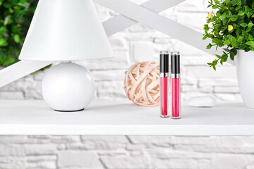 Two pink lip glosses on white rack next to a lamp, green plant in pot and round element of decor against brick wall. Close up, copy space