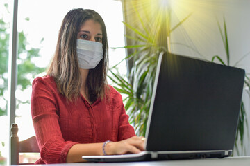 Young woman wearing a protective mask while she is working at home and attending at a meeting on her computer during the lockdown due to the covid