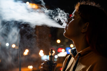 The girl smokes an electronic cigarette against the backdrop of the night city. Woman enjoying...