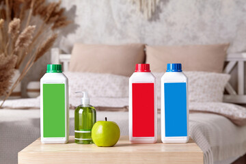 Three colorful plastic containers with detergents, pump bottle with soap and green apple on wooden bedside table in bedroom. Close up, copy space