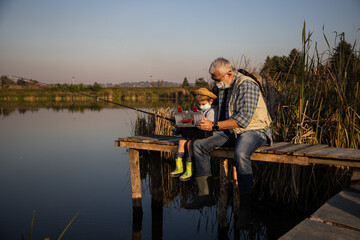 Senior man sitting on jetty fishing in lake with grandson under covid masks