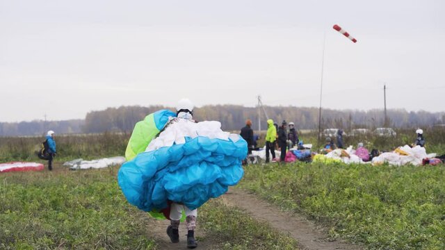 Man carries parachute. Action. Man carries multi-colored parachute after landing. Man in overalls drags parachute to gathering place in field. Extreme sport and skydiving