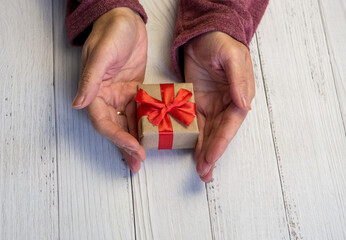 Women's hands hold a craft Christmas or new year decorated gift box. Light wooden background.