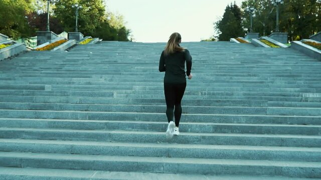 Young woman with ponytail wearing black sportswear running up long staircase in city in slow motion. Following shot of female athlete doing cardio training in the morning. Concept of fitness
