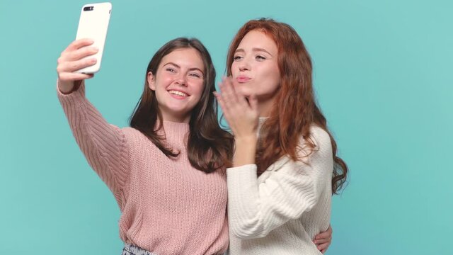 Two smiling young brunette redhead women friends 20s in casual pastel pink white sweaters hold using mobile cell phone doing selfie posing isolated on blue background studio . People lifestyle concept