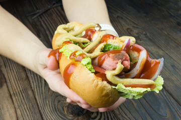 women's hands hold a delicious hot dog with a Bavarian sausage and the addition of fresh vegetables, ketchup and mustard.