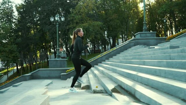 Fit woman with ponytail wearing black sportswear and white sneakers running up concrete staircase in park in slow motion, street lamps and trees on background. Tracking shot jogger exercising outside