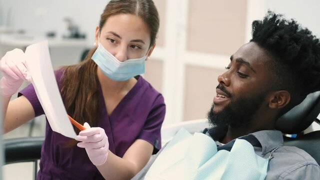 Young beautiful female dentist speaking with her handsome male dark skinned patient in dental clinic, showing xray image of his teeth. Dental and oral care concept.