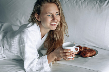 Beautiful blonde woman in white coat with white smile having breakfast in a hotel room. Woman is...