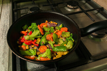 Lots of colorful vegetables are fried in a Frying pan on the kitchen stove. Carrots, capsicum, peppers, broccoli,  pisums, peas, corn and onions