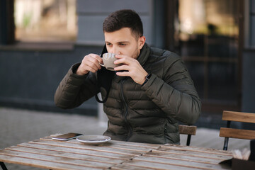 Man pulled off face mask for drinkig cappuccino on terrace. Man in cafe outdoors during quarantine. Covid-19