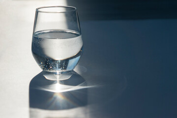 transparent glass of pure water on white table with ray through glass with reflection.  hard sunlight and shadows. Pure water concept