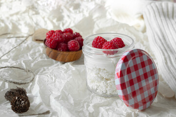 Red raspberries lie on cottage cheese with yogurt, sour cream in a jar, on crumpled white paper. Pullover, cones.