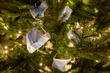 several mouth protection mask on a pine tree branch, lights in the background. Symbol for impact of covid-19 virus in christmas season 2020 and 2021
