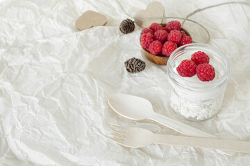 Red raspberries lie on cottage cheese with yogurt, sour cream in a jar, on crumpled white paper. Cones, paper hearts.