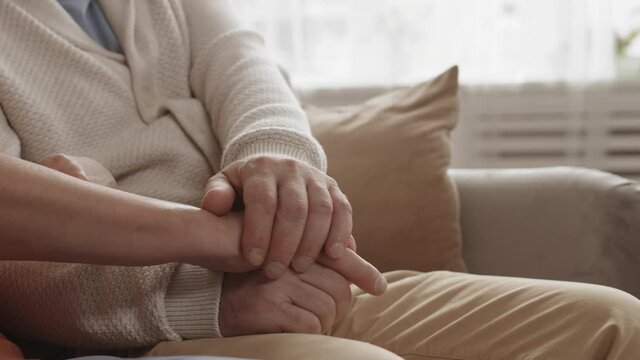 Close up of unrecognizable old man and woman sitting together on couch in living room and holding hands of each other