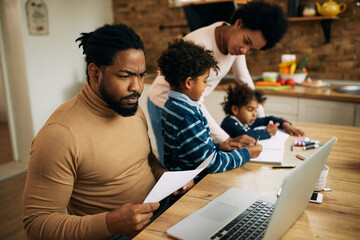 Black father working on laptop while mother is homeschooling their kids.