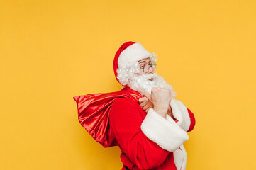 Fototapeta na wymiar Joyful and funny Santa Claus on a yellow background is holding a red sack loaded with presents and is going to bring the gifts to the kids.