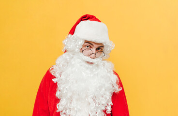 Fototapeta na wymiar Closeup portrait of a Santa Claus standing at the studio on a yellow background, looking at the camera with an interested expression on his face with eyeglasses on his nose.
