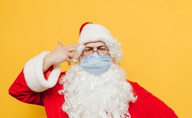 Funny portrait of Santa Claus wearing a disposable face mask, touching his head with two fingers imitating a shot gesture. Santa is feeling stressed during corona virus pandemic.