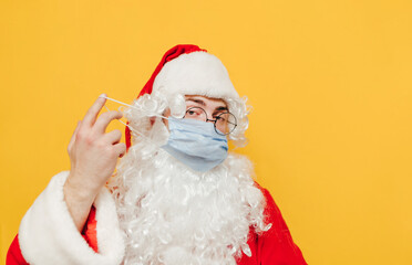 Closeup portrait of Santa Claus taking off his protective face mask, standing at the studio on a yellow background, looking at the camera.