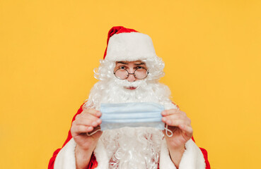 Fototapeta na wymiar Skeptical Santa Claus is holding a protective face mask, doubting that it will help during covid pandemic. Portrait of Santa with disposable mask on a yellow background.