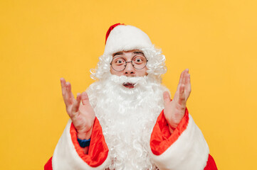 Fototapeta na wymiar Surprised Santa Claus is raising his hands up, looking at the camera with his eyes wide open, smiling, feeling astonished, posing on a yellow background.