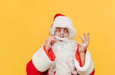 Attractive Santa Claus is talking with someone via mobile phone, having a discussion, gesticulating, raising his hand, trying to explain. Santa is standing at the studio on a yellow background.
