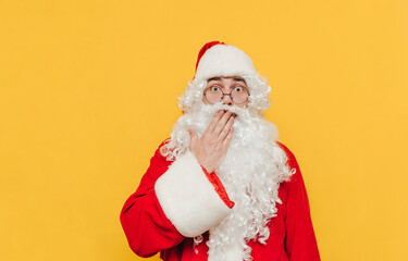 Surprised Santa Claus is touching his mouth with his hand showing an amazement gesture, feeling shocked, looking at the camera with his scared eyes wide open.