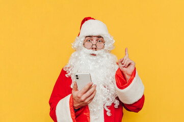 Cheerful Santa Claus with smartphone on a yellow background is having an idea, pointing his finger up, looking at the camera with his eyes wide open.