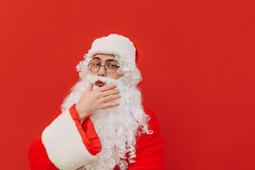 Santa Claus in fancy eyeglasses and a red costume is feeling thoughtful and touching his beard with his hand. Pensive Santa is looking at the camera, posing on a red background.