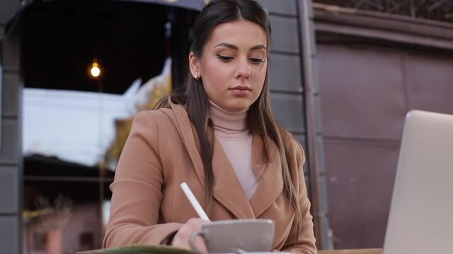 Young business woman sitting on terrace by cafe with laptop. Woman dring coffee and working outdoors. Female use phone and notebook