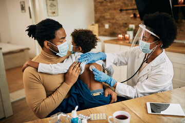 Black female doctor examining small boy with a stethoscope during home visit due to COVID-19...