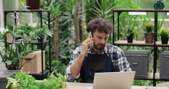 Young handsome male florist in apron sitting at desk in flower shop and typing on laptop while calling on cellphone. Joyful Caucasian man worker speaking on smartphone in floral center. Work concept