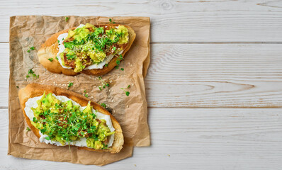 Two avocado sandwiches, tomato, microgreens and sesame seeds on baguette toast with cream cheese on a paper lining, white wooden table. Top view with copy space. Vegetarian toast
