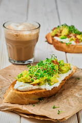 Close-up of avocado sandwich with cream cheese, microgreens and sesame seeds on paper lining. Glass with cappuccino, selective focus. Breakfast concept. Vegetarian toast