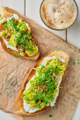 Avocado sandwich with cream cheese, microgreens and sesame seeds on paper lining. Top view. Cup with cappuccino, selective focus. Breakfast or brunch concept. Vegetarian toast
