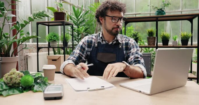 Portrait of handsome man in glasses and apron looking at laptop and writing down in planner while sitting in cozy floral shop. Caucasian young male florist flower store owner working. Business concept