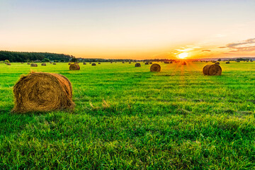 Scenic view at beautiful sunset in a green shiny field with hay stacks, cloudy sky, golden sun...